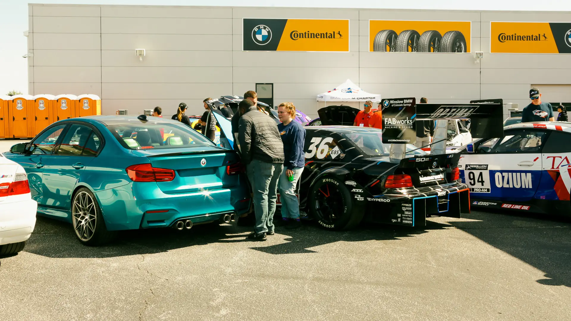 Teal BMW M3 beside race cars at a car show under Continental banners