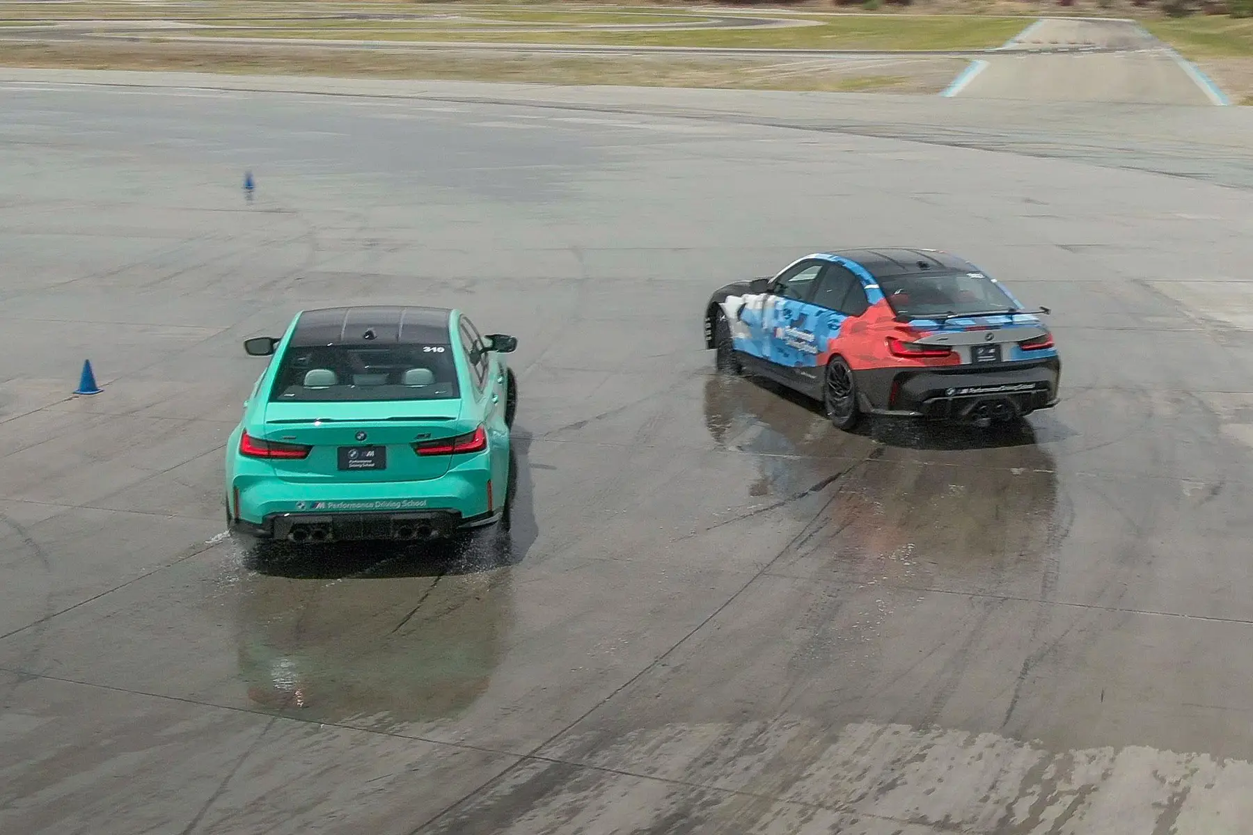Mint green BMW M3 and a liveried M3 drifting on the wet pad, seen from above