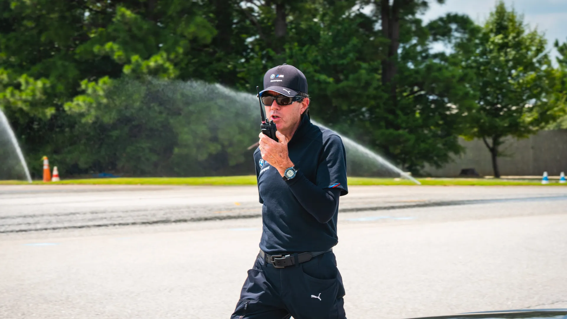 Instructor speaking into a handheld radio as sprinklers water the skid pad behind him