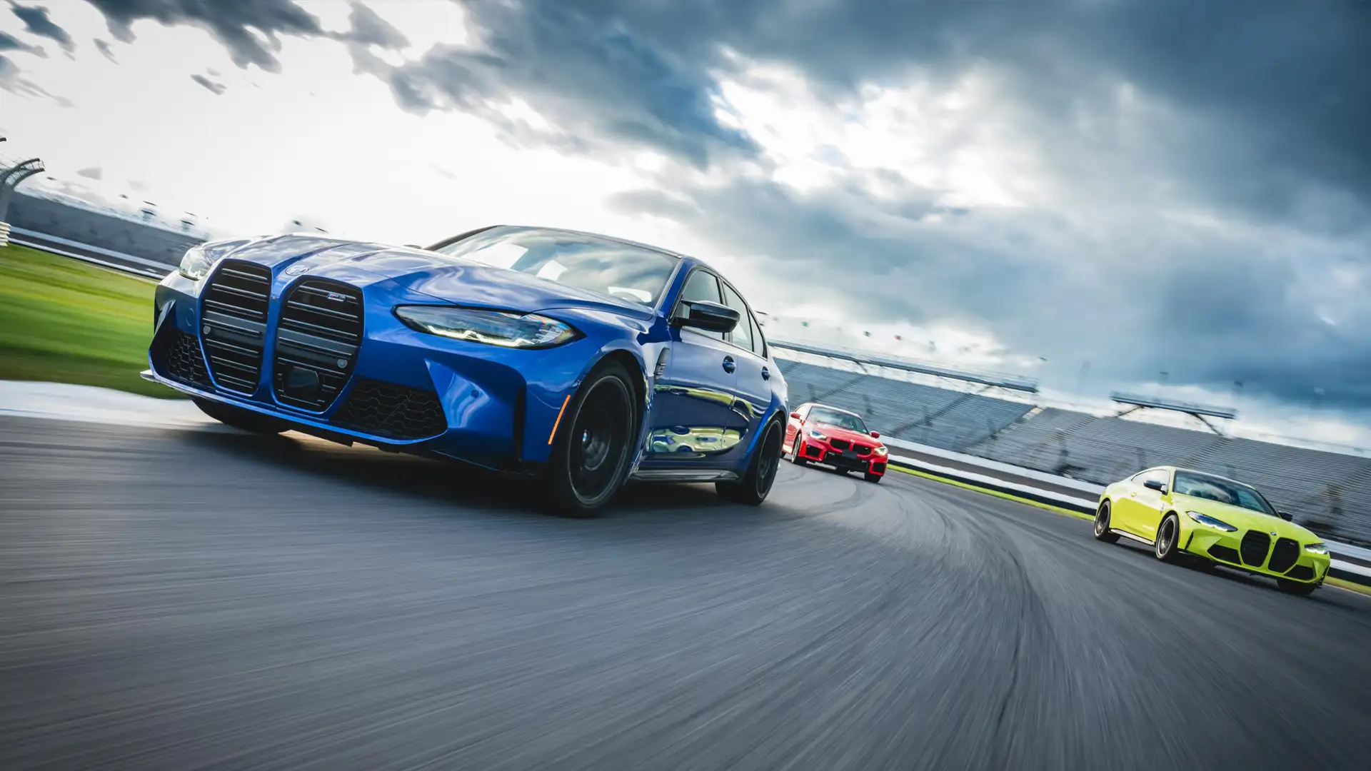 Blue BMW M3 leading a red M2 and a lime green M4 through a banked corner