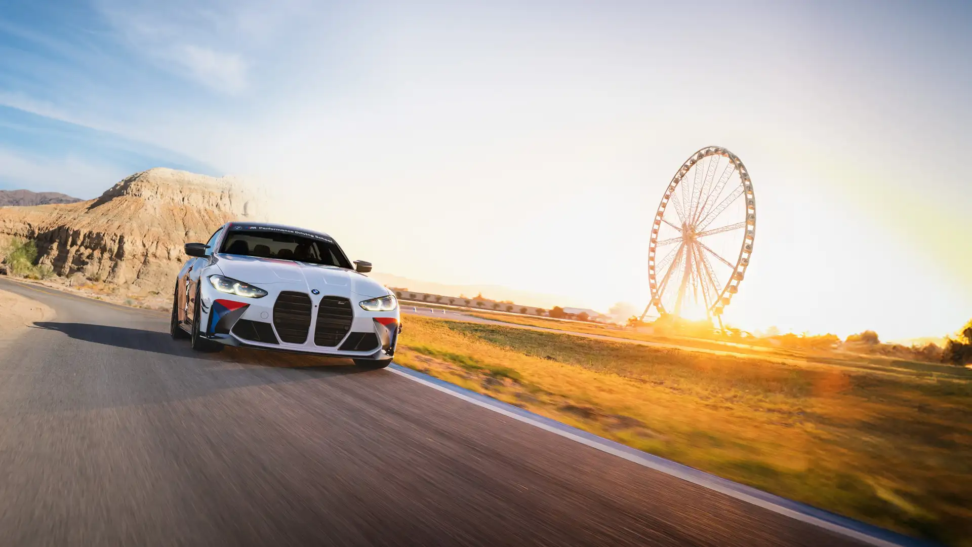 White BMW M4 on a desert road with a Ferris wheel silhouetted at sunset
