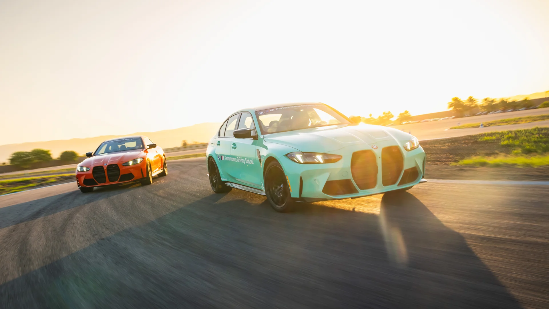 Mint green BMW M3 and orange BMW M4 cornering on the desert circuit at sunset