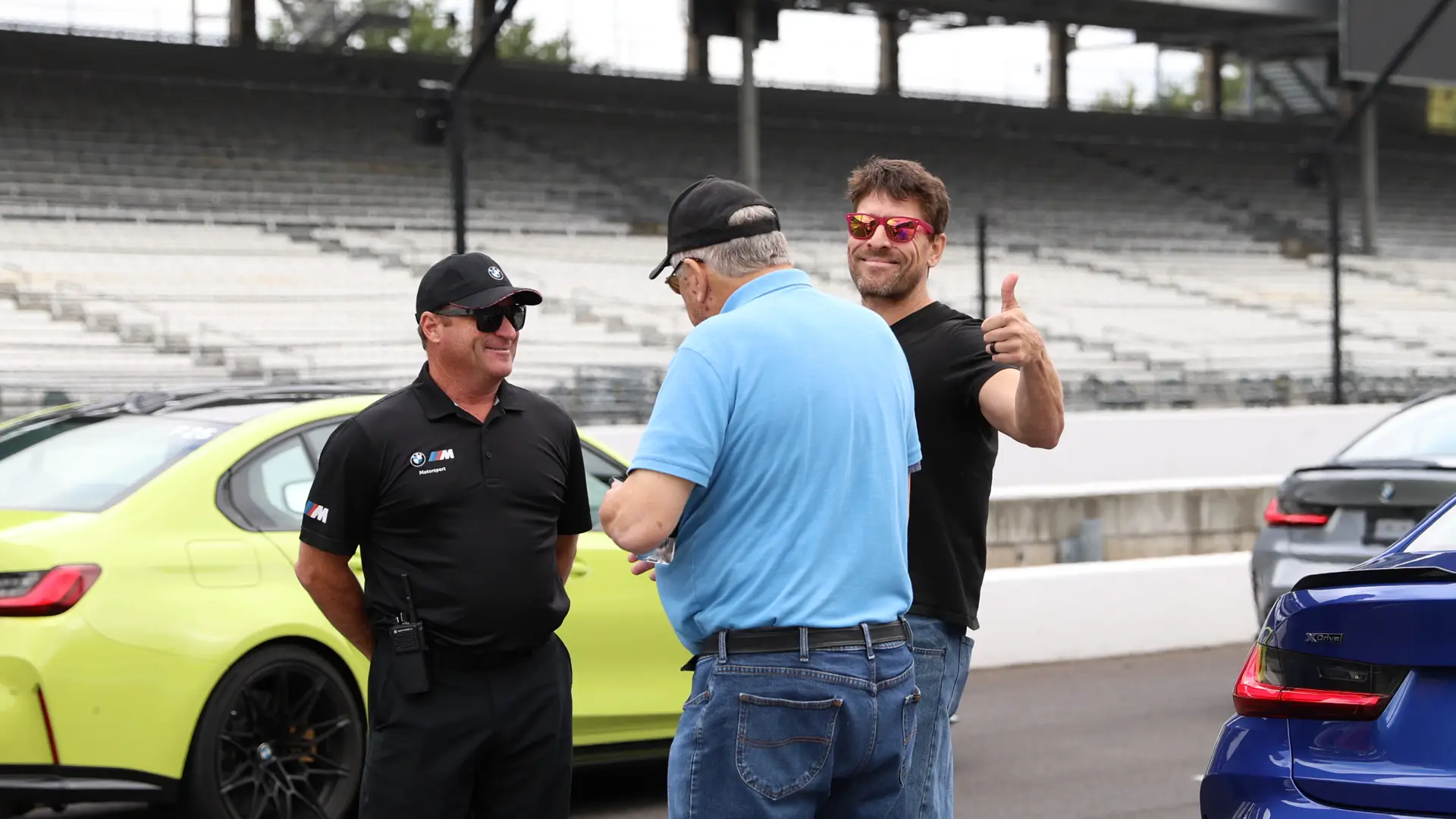 Participants talking beside BMW M cars in the grandstands at Indianapolis, one giving a thumbs up