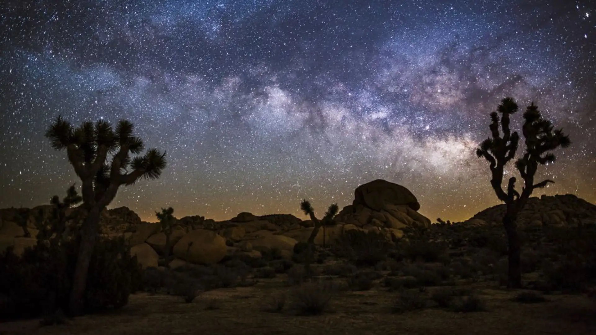 The Milky Way arching over Joshua trees and desert boulders at night