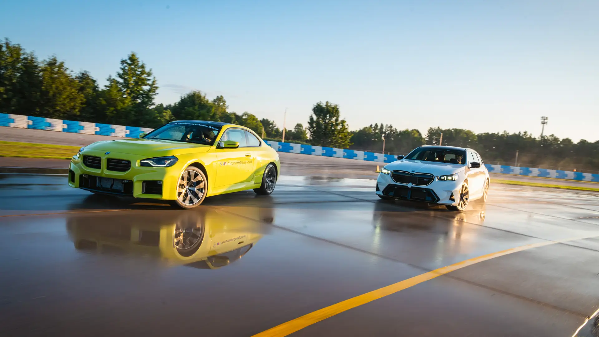 Lime green BMW M2 and a white M5 on the polished wet skid pad at sunset