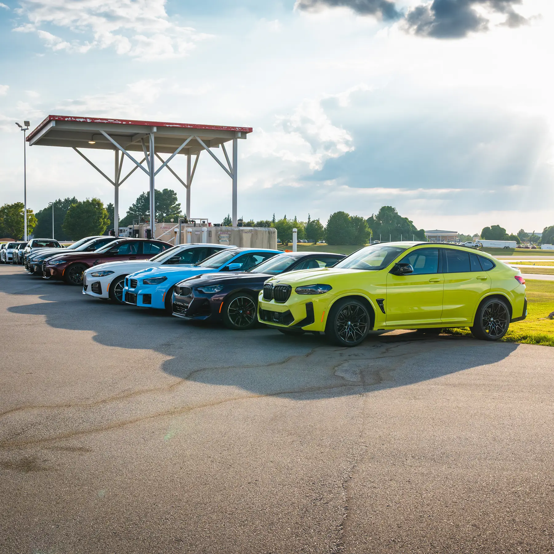 Line-up of BMW M cars led by a lime green X4 M beside the trackside canopy
