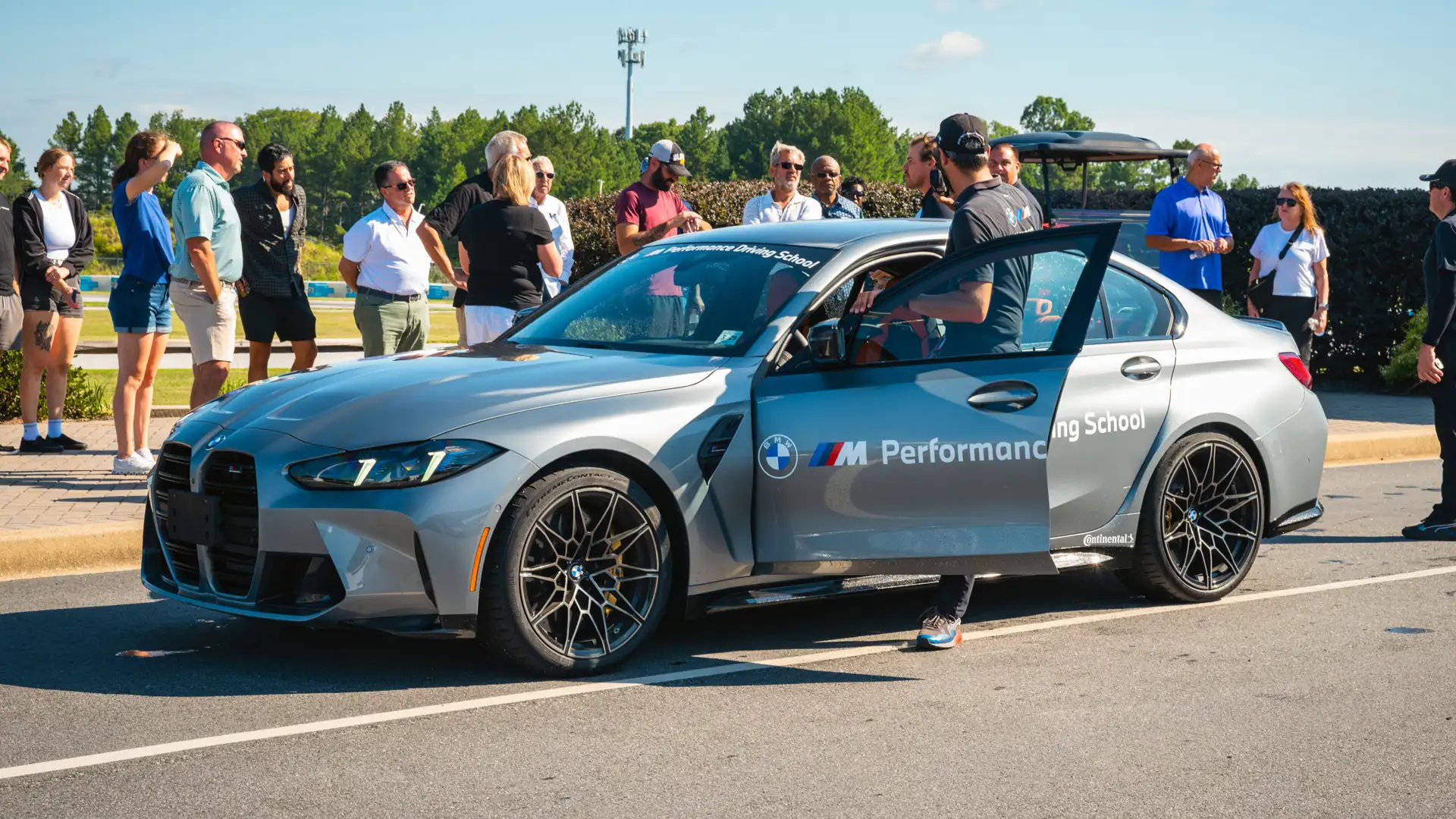 Group gathered around a grey BMW M3 as an instructor opens the driver's door