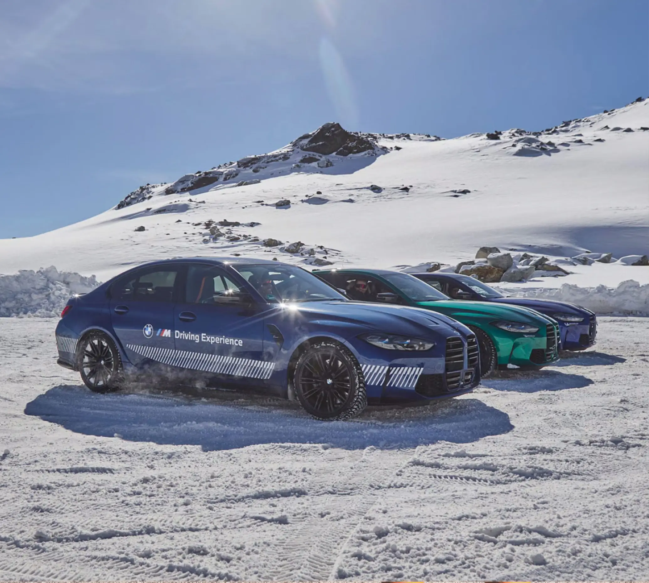 Line of BMW M Driving Experience cars parked on the snow below a mountain ridge