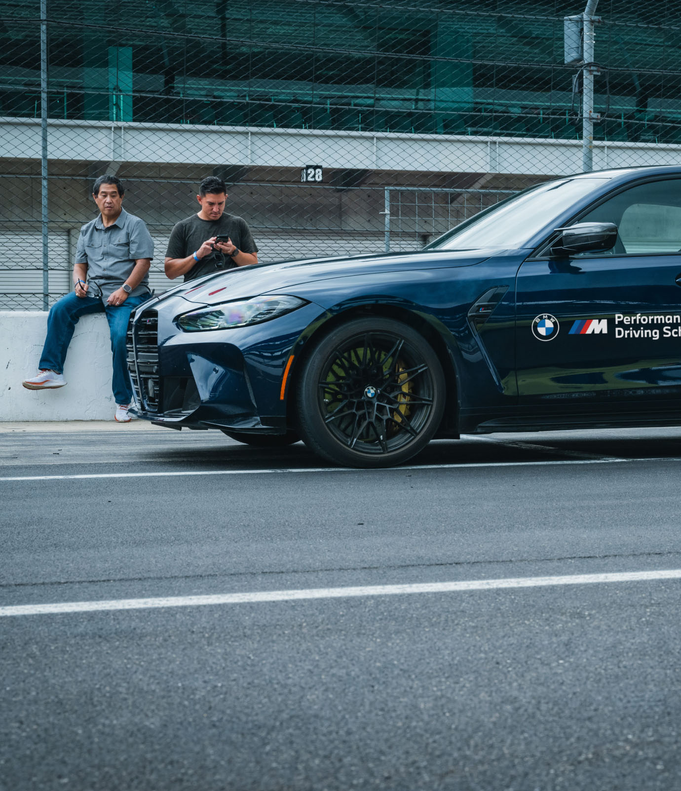 Two participants beside a dark blue BMW M4 in the pit lane at Indianapolis Motor Speedway